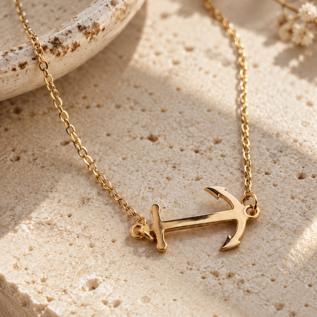 A gold anchor pendant necklace on a delicate gold chain, styled on a textured beige stone surface with warm natural shadows.