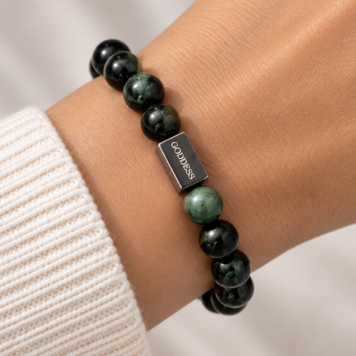 A close-up of a beaded bracelet worn on a wrist, featuring dark green and black marbled beads, a silver rectangular charm engraved with the word 'GODDESS', and a cream ribbed knit cuff.