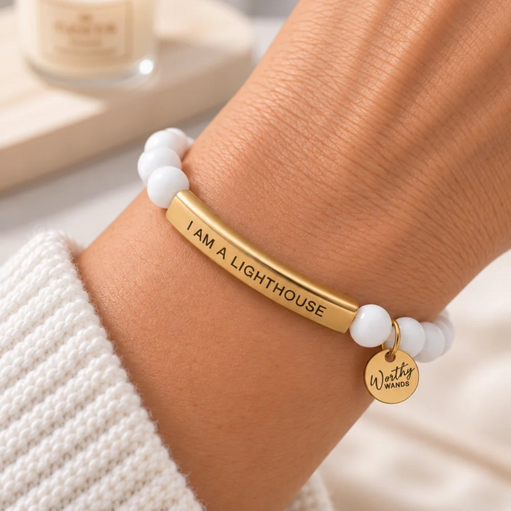 a gold bar bracelet engraved with 'I AM A LIGHTHOUSE' and a small round charm reading 'Worthy Wands' on white beads