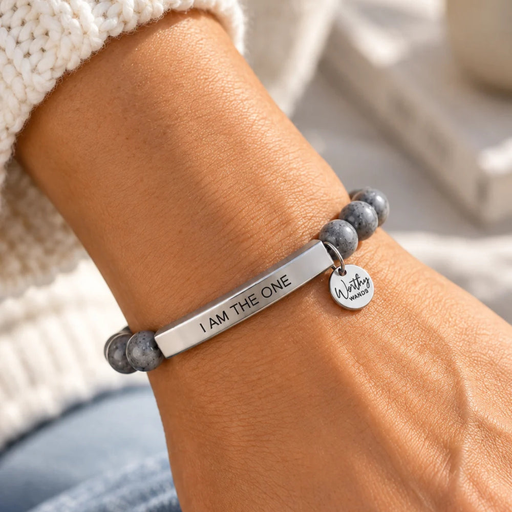 A close-up of a person's wrist wearing a silver bar bracelet engraved with 'I AM THE ONE' The bracelet is styled with gray beaded elements and a chunky white knit sweater cuff is visible.