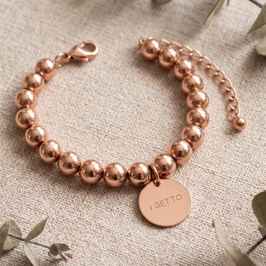 Rose gold beaded bracelet with a round engraved disc charm reading 'I GET TO', displayed on a textured beige fabric background with eucalyptus leaves.